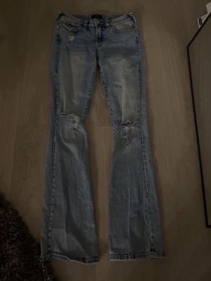 Ljusblå bootcut jeans från True Religion - Säljer ett par ljusblå bootcut jeans från True Religion med slitningar och hål på knäna. Jeansen har klassiska bakfickor med broderad logga och silverfärgade knappar. Även low waist.