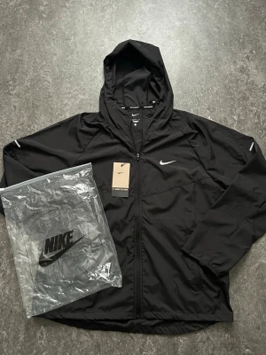 Svart Nike vindjacka med huva - Svart vindjacka från Nike med huva och dragkedja framtill. Jackan har två sidofickor, reflekterande detaljer på ärmarna och klassisk Nike-logga på bröstet. Tillverkad i lätt och vattenavvisande material, perfekt för blåsiga dagar.