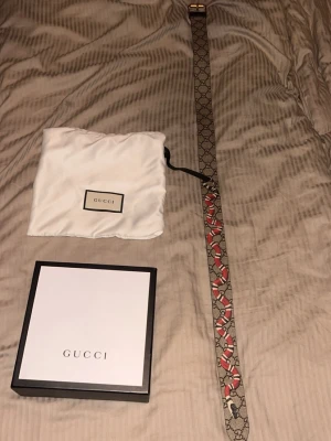 Gucci bälte med orm och GG-mönster - Snyggt Gucci bälte i skinn med klassiskt GG-mönster och en röd orm som detalj längs bandet. Bältet har ett guldfärgat spänne och levereras med originalpåse och box. Perfekt accessoar för att lyfta din outfit. Bältet säljs inte längre så svårt o få tag i