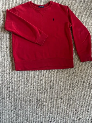 Röd sweatshirt från Polo Ralph Lauren barn - Röd sweatshirt från Polo Ralph Lauren med rund halsringning och broderad logga på bröstet. Tröjan har långa ärmar och ribbade muddar vid ärmslut och nederkant. Perfekt för en avslappnad och stilren look. Den har små fläckar som jag har tagit bild på.