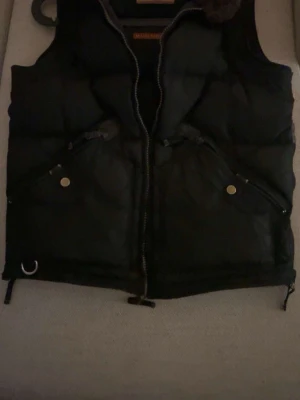 Svart dunväst från Parajumpers - Snygg svart dunväst från Parajumpers med dragkedja framtill, två fickor med tryckknappar och detaljer i metall. Västen har en klassisk puffer-design och hög krage. Perfekt för lager-på-lager och streetstyle. PRIS KAN DISKUTERAS VID SNABB ARFFÄR!