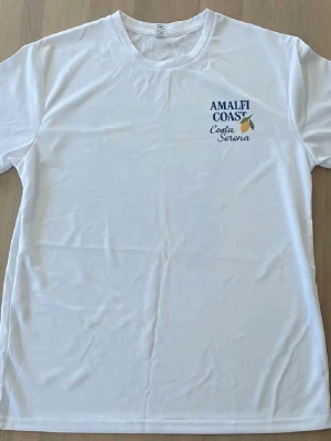Vit Amalfi Coast t-shirt med tryck - Säljer en vit t-shirt med färgglatt Amalfi Coast-tryck på ryggen och mindre tryck framtill. Motivet visar citroner, hav och en flaska vin, samt texten Costa Serena. T-shirten har rund hals och korta ärmar, perfekt för sommarens äventyr.