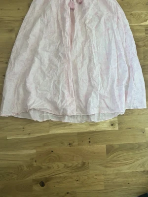 Ljusrosa långkjol med knyt - Ljusrosa långkjol med diskret mönster och elastisk midja. Kjolen har ett knytband med tofsar framtill och är tillverkad i ett lätt bomullsmaterial. Perfekt för dig som gillar en luftig och avslappnad stil.