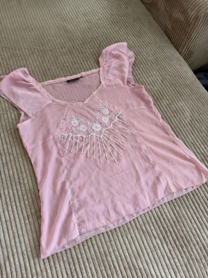 Vintage Ljusrosa blus med pärlor och broderi - Supersöt ljusrosa blus med v-ringning och korta ärmar. Framsidan har detaljerat vitt blombroderi och pärlor som ger en unik look. Tillverkad i ett tunt, mjukt tyg med spetsdetaljer vid halsen. Perfekt för dig som gillar romantisk och feminin stil.