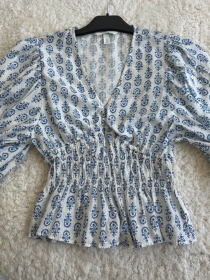 Vit och blå blus med v-ringning - Söt blus i vitt med blått blommigt mönster och v-ringad hals. Modellen har puffiga långärmar och smockad midja som ger en feminin siluett. Perfekt till jeans eller kjol för en trendig look.