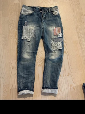 Patchade boyfriendjeans med spets - Unika blå boyfriendjeans med flera coola lappar i spets, brodyr och randigt tyg. Jeansen har avslappnad passform, slitningar och spetsdetaljer vid benslut. Perfekta för dig som gillar en personlig och kreativ stil.