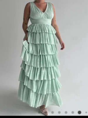 Ljusgrön volangklänning med v-ringning - Superfin långklänning i ljusgrönt med flera lager volanger och v-ringad hals. Klänningen har breda axelband och markerad midja, vilket ger en elegant och flowy look. Perfekt för bal eller student. Materialet känns lätt och luftigt. Använd 1 gång och är perfekt på mig som är 175, original pris är 1800 och säljs inte längre