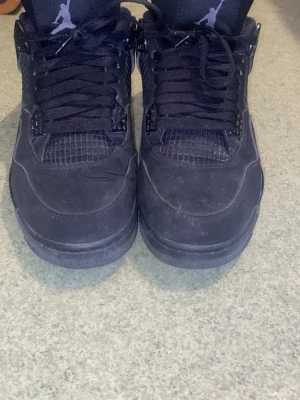 Svarta Air Jordan 4 Retro sneakers - Säljer ett par helsvarta Air Jordan 4 Retro sneakers med klassisk Jumpman-logga på plösen och hälen. Skorna har meshdetaljer, robust sula och coola plastdetaljer vid snörningen. Tillverkade i mocka och textil för en stilren look.