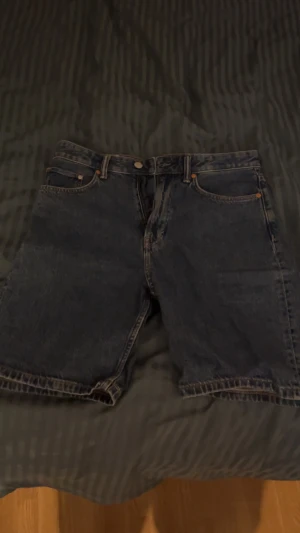 Mörkblå jorts med 5 fickor - Säljer ett par mörkblå jeansshorts i klassisk modell med fem fickor och knappgylf. Shortsen har raka ben och slitstark denimkänsla, perfekta för en avslappnad stil. Passar dig som gillar tidlös och enkel design.