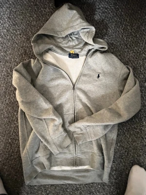  Polo Ralph Lauren Zip Up - Snygg grå hoodie från Polo Ralph Lauren med dragkedja framtill och klassisk huva. Ikoniska lilla loggan broderad på bröstet. Tillverkad i mjukt bomullsmaterial och har två fickor framtill. Perfekt för en chill och avslappnad stil.