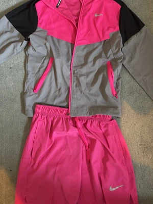 Nike miler tracksuit - Nike Running träningsset med jacka och shorts. Jackan är grå och rosa med svarta axelpartier, dragkedja och två fickor med rosa detaljer. Shortsen är helrosa med resår och dragsko i midjan. Båda plaggen har Nike-logga och är tillverkade i lätt, ventilerande material.
