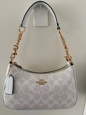 Vit axelremsväska från Coach - Snygg vit handväska från Coach med klassiskt monogrammönster i beige och vitt. Väskan har guldfärgade kedjedetaljer och dragkedja upptill. Axelremmen är i vitt läder med guldfärgade länkar. Perfekt för dig som gillar stilrena accessoarer.