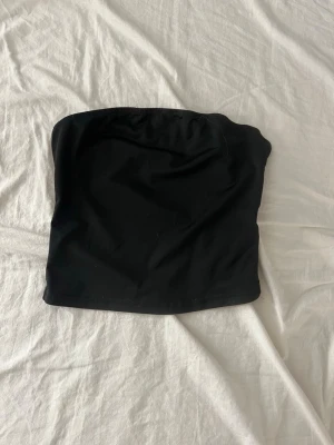 Svart bandeau tubtopp från SHEIN - Svart bandeau tubtopp från SHEIN i storlek XS. Enkel och stilren design utan axelband, perfekt att matcha med olika outfits. Materialet är syntet och toppen har en tight passform som sitter snyggt på kroppen.
