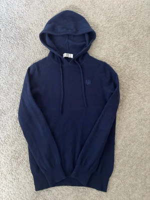 Mörkblå hoodie från BVALDI - Mörkblå hoodie från BVALDI med broderad logga på bröstet. Tröjan har dragsko i huvan och är tillverkad i ett mjukt, stickat material. Långa ärmar och ribbade muddar ger en skön passform. 