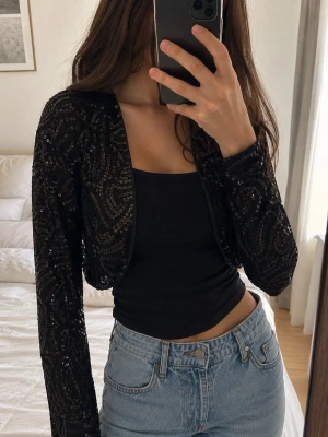 Svart paljettbolero med lång ärm - Snygg svart bolero med långa ärmar, täckt av glittrande paljetter i mönster. Kort modell som passar perfekt över linne eller klänning. Ger en cool och festlig vibe till din outfit.