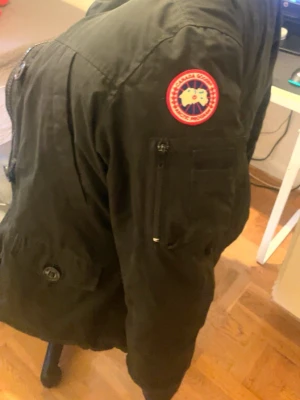 Svart dunjacka från Canada Goose - Snygg svart dunjacka från Canada Goose med klassisk patch på ärmen och stor huva med pälskant. Jackan har dragkedja och knappar framtill samt flera fickor. Perfekt för kalla vinterdagar och riktigt varm.