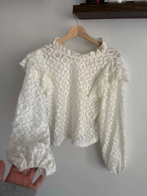 Vit spetsblus med volangdetaljer - Superfin vit blus i spets med volangdetaljer vid axlar och hals. Blusen har långärmade puffärmar och är tillverkad i ett genombrutet spetsmaterial som ger en romantisk känsla. Perfekt att styla med jeans eller kjol för en trendig look.
