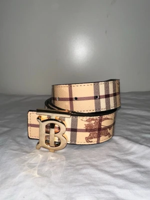 Beige rutigt Burberry bälte - Snyggt bälte från Burberry i klassiskt beige och brunt rutigt mönster. Bältet har ett stort guldfärgat B-spänne och är gjort i skinn. Perfekt accessoar för att lyfta din outfit med en lyxig touch.