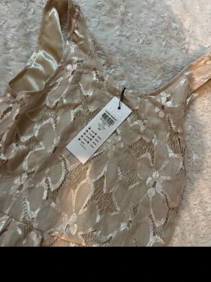 Beige spetsklänning med blommönster - Söt beige klänning med blommig spets och ärmlös design. Klänningen har en lätt glansig underton och markerad midja. Perfekt för dig som gillar romantiska detaljer och en feminin look.