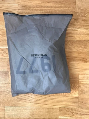 Grå Essentials Fear of God hoodie - Snygg grå hoodie från Essentials Fear of God med svart tryck '1977' på bröstet. Klassisk passform med huva och mjukt material som känns skönt mot huden. Perfekt för dig som gillar streetwear och vill ha en stilren look.