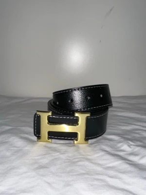 Svart skärp med guldspänne - Stilrent svart skärp med ett ikoniskt H-format spänne i guldmetall. Skärpet har vita kontrastsömmar längs kanterna och är tillverkat i slätt skinn. Perfekt accessoar för att lyfta din outfit.