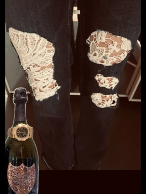 Mörkblå bootcut jeans med spets - Unika mörkblå bootcut jeans med stora slitningar framtill som är täckta med vit spets. Jeansen har klassisk femficksmodell och kontrastsömmar. Perfekta för dig som vill sticka ut med en cool och kreativ stil.