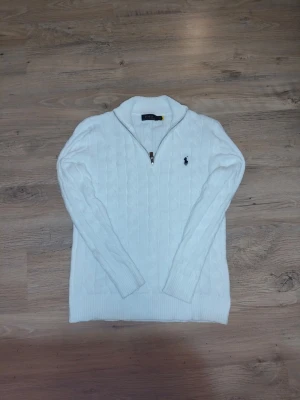 Vit kabelstickad tröja Polo Ralph Lauren - Vit kabelstickad tröja från Polo Ralph Lauren med halv dragkedja och broderad logga på bröstet. Tröjan har lång ärm, ribbstickade muddar och klassisk passform. Perfekt för lager-på-lager och snygg till jeans.