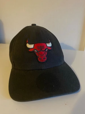 Svart Chicago Bulls keps - Säljer en svart keps med Chicago Bulls-logga broderad framtill. Klassisk böjd skärm och justerbar passform bak. Tillverkad i slitstark bomull med tydlig röd och vit detalj på loggan. Perfekt för dig som gillar basket och streetwear.
