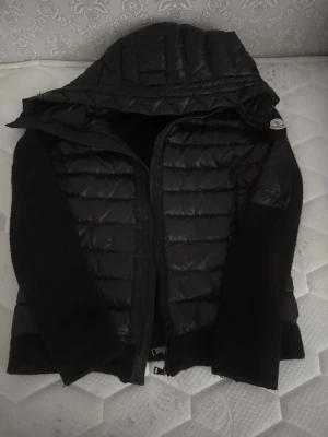 Svart pufferjacka med huva Moncler - Snygg svart pufferjacka från Moncler med glansig vadderad front och huva. Ärmarna är i mjukt stickat material och jackan har dragkedja framtill samt fickor med dragkedja. Perfekt för kalla vinterdagar och riktigt trendig look. Den är äkta du kan få mer bilder om du vill 