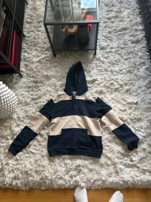 Tommy Hilfiger hoodie i marinblått och beige - Snygg hoodie med breda marinblå och beige ränder. Tröjan har huva, känguruficka framtill och ribbade muddar vid ärmslut och nederkant. Perfekt för en avslappnad och trendig stil.