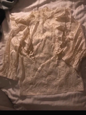 Krämvit spetsblus med volanger - Romantisk krämvit blus i spets med volangdetaljer längs bröstet och axlarna. Blusen har hög krage med smock och långa ärmar med smockade muddar. Perfekt för dig som gillar en feminin och vintageinspirerad stil.