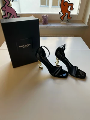 Svarta Saint Laurent sandaler med YSL-klack - Säljer ett par svarta klackskor från Yves Saint Laurent i lackat läder med öppen tå och ett smalt band över foten. Klacken är i guld och formad som YSL-loggan, vilket ger en snygg och lyxig vibe. Justerbart ankelband för perfekt passform. Riktigt statement piece för dig som gillar designer. Storlek 39, säljer dem för 2500kr och skickar så snabbt jag kan!