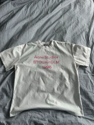 Vit Acne Studios t-shirt med tryck - Vit t-shirt från Acne Studios med rosa texttryck 'Acne Studios STOCKHOLM 1996' på bröstet. Klassisk passform med rund hals och korta ärmar. Perfekt för en clean och stilren look.