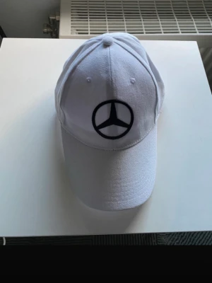 Vit keps med Mercedes-Benz logo - Snygg vit keps med klassisk böjd skärm och broderad svart Mercedes-Benz logotyp framtill. Kepsen har justerbar rem baktill och är tillverkad i bomullstyg. Perfekt accessoar för dig som gillar stilrena detaljer och bilar.