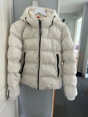 Superfin vit pufferjacka från superdry 🤍 - Varm och skön oversized modell med hög krage och svarta detaljer. Perfekt till höst/vinter. Använd, finns mindre märken/skrap på ärmen och några små fläckar (se bild), därav lägre pris. Annars fint skick. Skriv vid frågor eller fler bilder 💌
