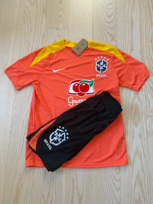 Brasilien fotbollsträningsset Nike - Snyggt träningsset från Nike med Brasilien-tryck. Tröjan är orange med gula axelpartier och sponsorlogga, medan shortsen är svarta med orange detaljer och Brasilien-emblem. Perfekt för fotbollsträning eller match.