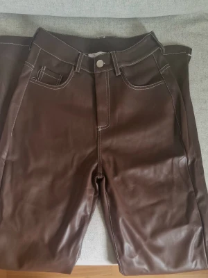 Bruna skinnbyxor - Snygga bruna byxor i skinn med vita kontrastsömmar och klassisk femficksdesign. Perfekta för dig som vill sticka ut med en trendig look. Byxorna har normal passform och raka ben.