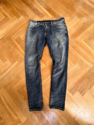 Blå slim fit jeans från Nudie - Snygga blå slim fit jeans från Nudie med klassisk femficksdesign och slitna detaljer. Jeansen har orangea sömmar och subtila slitningar framtill och baktill. Tillverkade i bomull med en autentisk tvättad look. W:32 L:32. Modell Skinny Lin