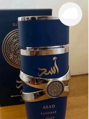 Asad Zanzibar parfym blå och silver - Snygg parfymflaska från Lattafa i djupblå färg med silvriga detaljer och orientaliskt mönster. Flaskan har en modern, rund form och pryds av eleganta arabiska tecken samt texten Asad Zanzibar. Perfekt för dig som gillar exklusiva dofter.