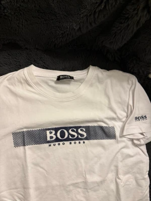 Vit Hugo Boss t-shirt med tryck - Snygg vit t-shirt från Hugo Boss med stort blått logotryck på bröstet och mindre logga på ärmen. Klassisk passform och rund halsringning. Tillverkad i mjuk bomull som känns skön mot huden. Perfekt för en clean och stilren look.