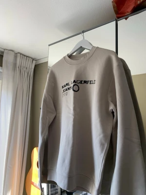 Beige sweatshirt Karl Lagerfeld  - Stilren beige sweatshirt från Karl Lagerfeld Jeans med svart tryck framtill. Tröjan har rund halsringning och långa ärmar, perfekt för en avslappnad och trendig look.