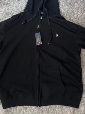 Svart hoodie från Polo Ralph Lauren - Svart hoodie med dragkedja från Polo Ralph Lauren. Klassisk design med huva, dragsnören och fickor framtill. Liten vit logga broderad på bröstet. Perfekt för en chill och stilren look.