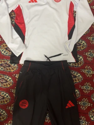 Bayern München träningsset Adidas - Snyggt träningsset från Adidas med Bayern München-logga. Tröjan är vit med röda och svarta detaljer, långärmad och har dragkedja vid halsen. Byxorna är svarta med röda detaljer och loggor, samt dragsko i midjan. Perfekt för fotbollsträning eller gym.