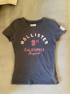 Mörkgrå Hollister t-shirt med tryck - Mörkgrå t-shirt från Hollister med rund hals och korta ärmar. Framsidan har tryck i vitt och rosa med texten 'Hollister 9th California Original'. Slimmad passform och mjukt bomullsmaterial. 