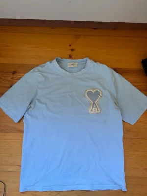 Ljusblå Ami Paris t-shirt med hjärtdetalj - Snygg ljusblå t-shirt från Ami Paris med korta ärmar och rund halsringning. På bröstet finns ett stort broderat hjärta med bokstaven A i vitt. Perfekt för en stilren och trendig look.