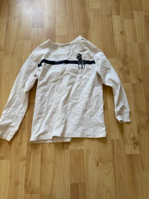 Vit långärmad t-shirt från Polo Ralph Lauren - Vit långärmad t-shirt från Polo Ralph Lauren med en marinblå rand över bröstet och stor logga på vänster sida. Siffran 3 i marinblått på höger ärm ger en sportig touch. Tillverkad i mjuk bomull och har normal passform.  Storlek S (8) kids