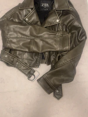Olivgrön skinnjacka från Zara - Cool olivgrön skinnjacka från Zara med klassisk bikerstil, silverfärgade dragkedjor och bälte i midjan. Jackan har breda slag, axelklaffar och detaljerade sömmar som ger en edgy look.