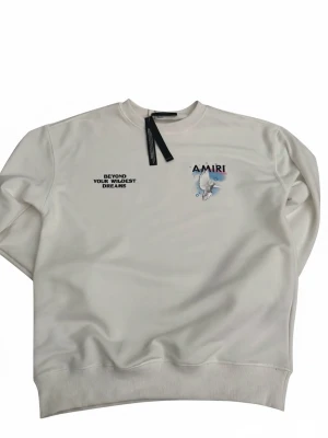 Vit sweatshirt från Amiri - Vit sweatshirt från Amiri med tryckt text ”BEYOND YOUR WILDEST DREAMS” och en duva tillsammans med Amiri-loggan på bröstet. Baksidan har också en duva med tryckt text ”Miracles Happen Everyday”. Köpt från Bloomingdale’s i Kuwait.