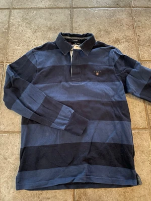 Randig rugbytröja från GANT - Snygg långärmad rugbytröja från GANT i marinblått och mörkblått med breda ränder. Klassisk krage och broderad logga på bröstet. Perfekt för en avslappnad och sportig stil.