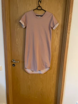Rosa t-shirtklänning från JDY - Enkel och stilren rosa t-shirtklänning från JDY i storlek XS. Klänningen har rund halsringning, korta ärmar med uppvik och rak passform. Perfekt för en avslappnad look och lätt att matcha med sneakers eller sandaler.